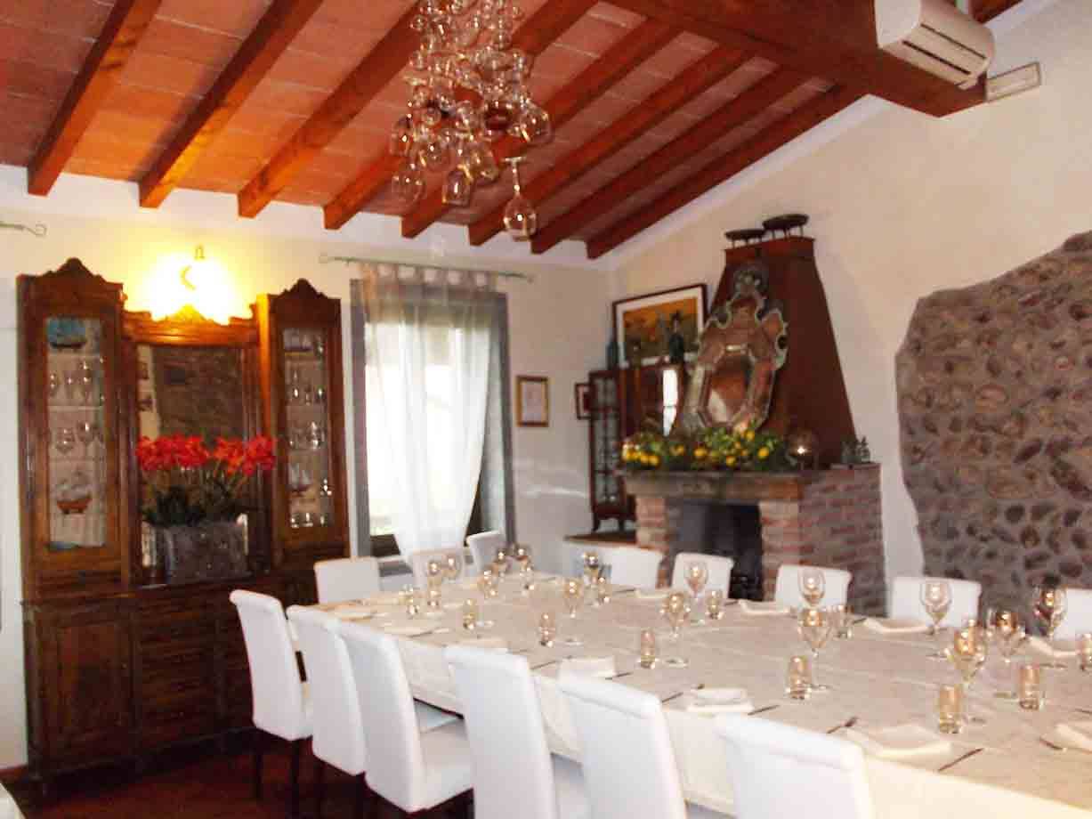 Sala da pranzo apparecchiata con tavolo lungo, sedie bianche, soffitto in legno e camino