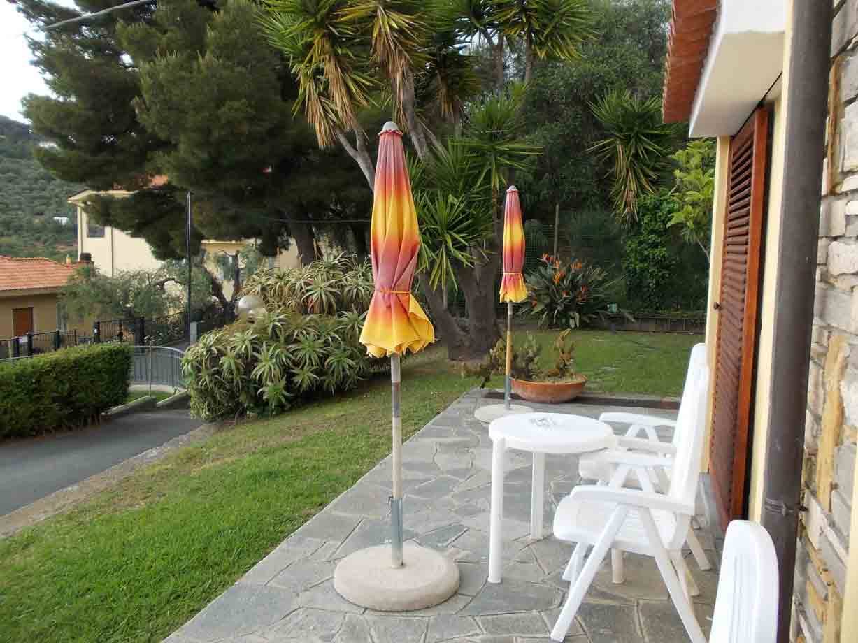 Terrazza con sedie e tavolo bianchi, ombrelloni chiusi tra yucca e siepi