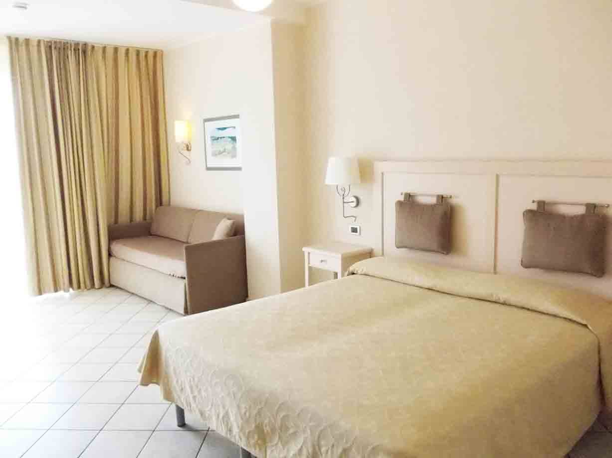 Camera d'albergo con letto matrimoniale, divano beige e tende chiare
