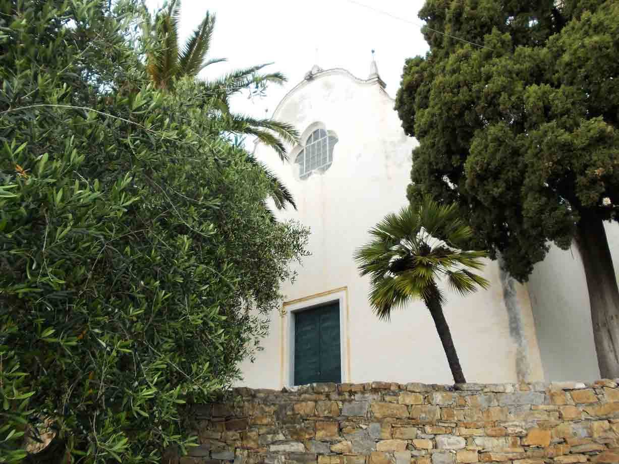 Chiesa bianca con portale verde tra palme, ulivi e muro di pietra