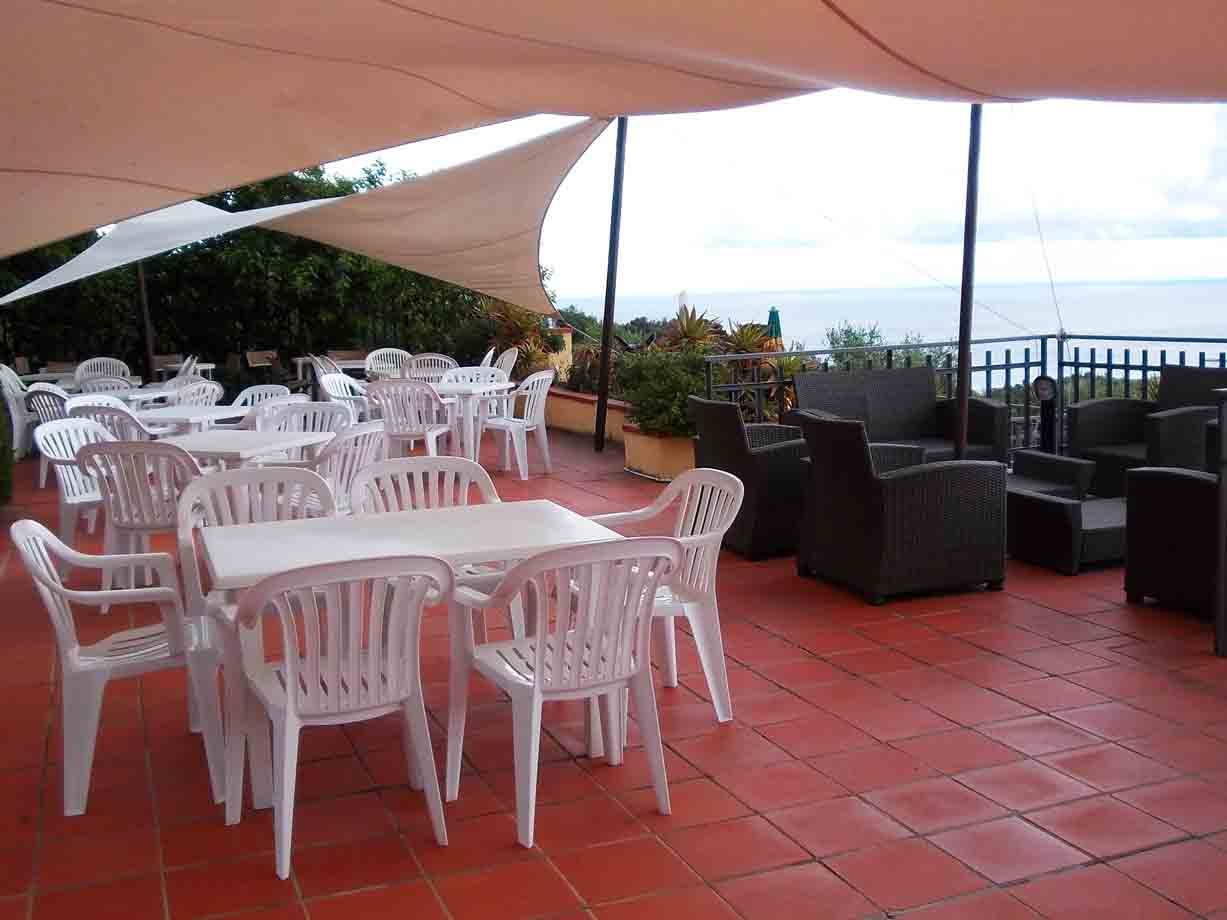 Terrazza con tavoli e sedie di plastica bianca sotto teli ombreggianti, davanti al mare