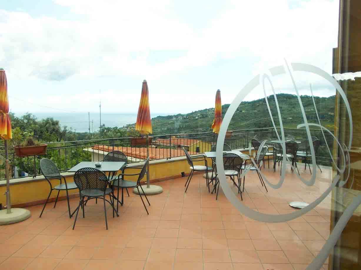 Terrazza con tavoli e sedie in ferro, ombrelloni chiusi, colline e mare sullo sfondo