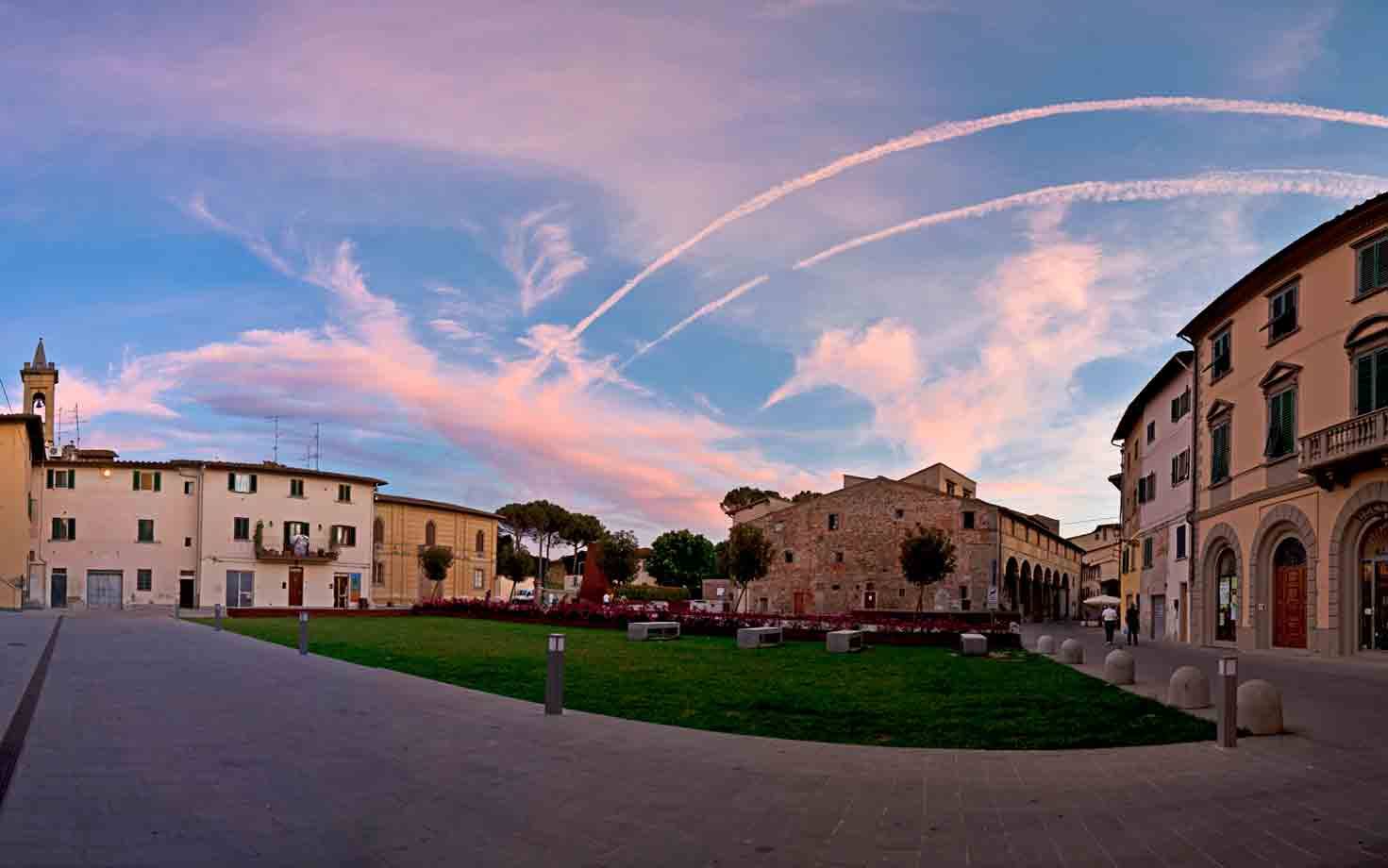 Piazzale con prato verde, edifici in pietra e cielo rosa striato di scie