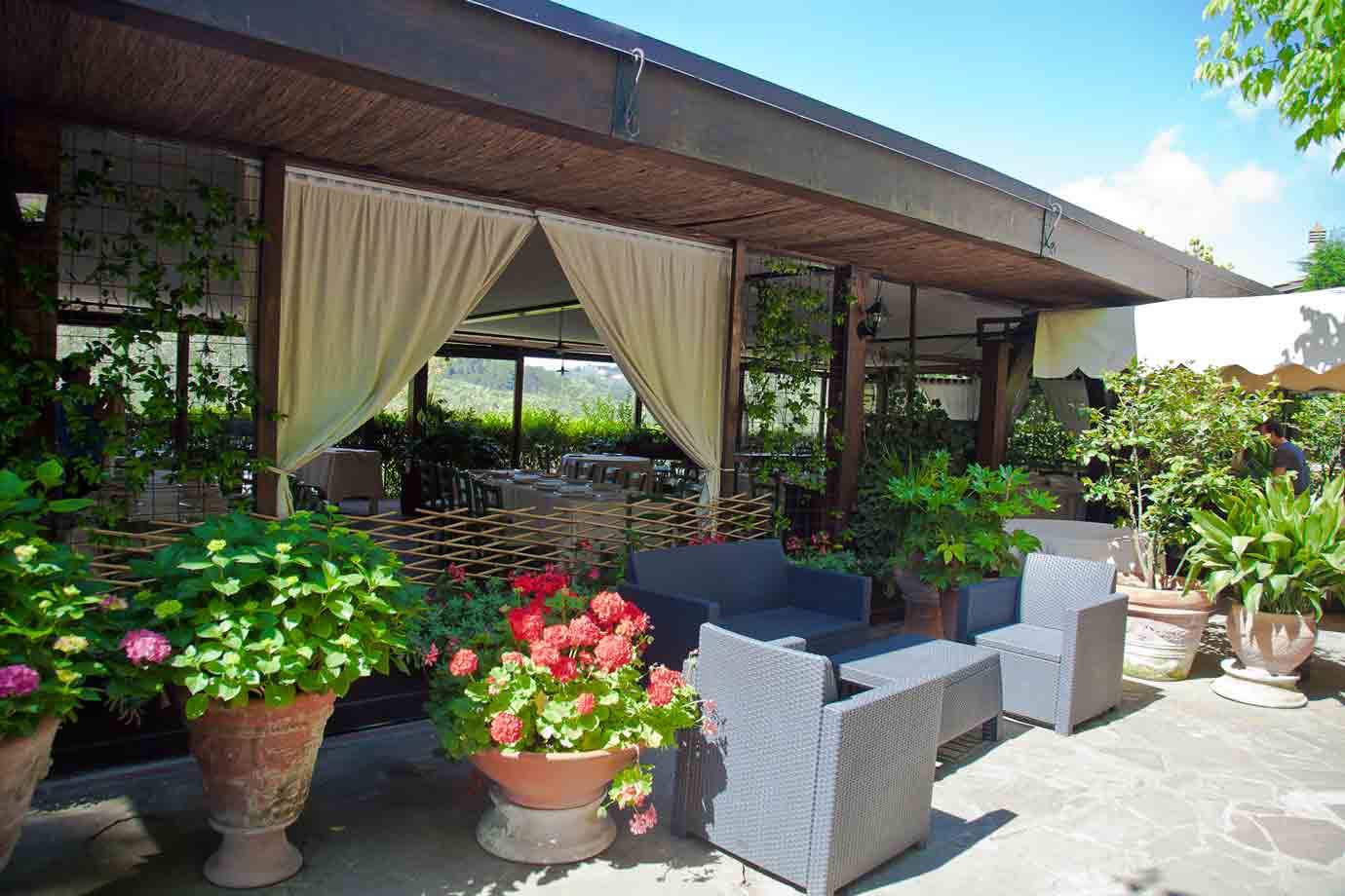 Terrazza ristorante con poltrone grigie, tende beige e vasi di gerani in fiore