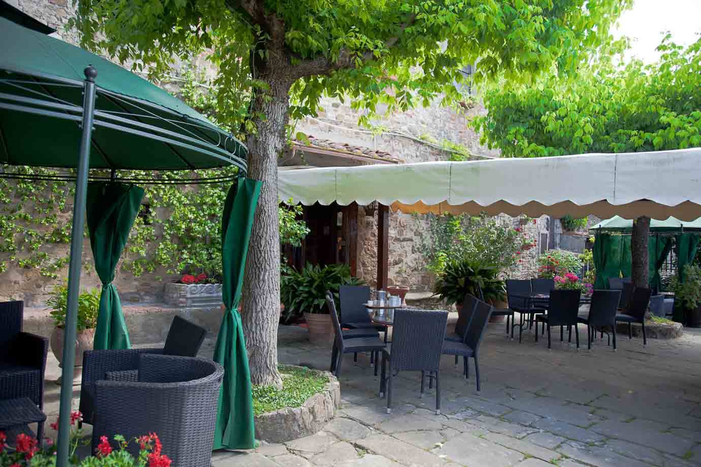 Tavoli vuoti sotto tende verdi e gazebo in pietra, cortile ombreggiato da alberi