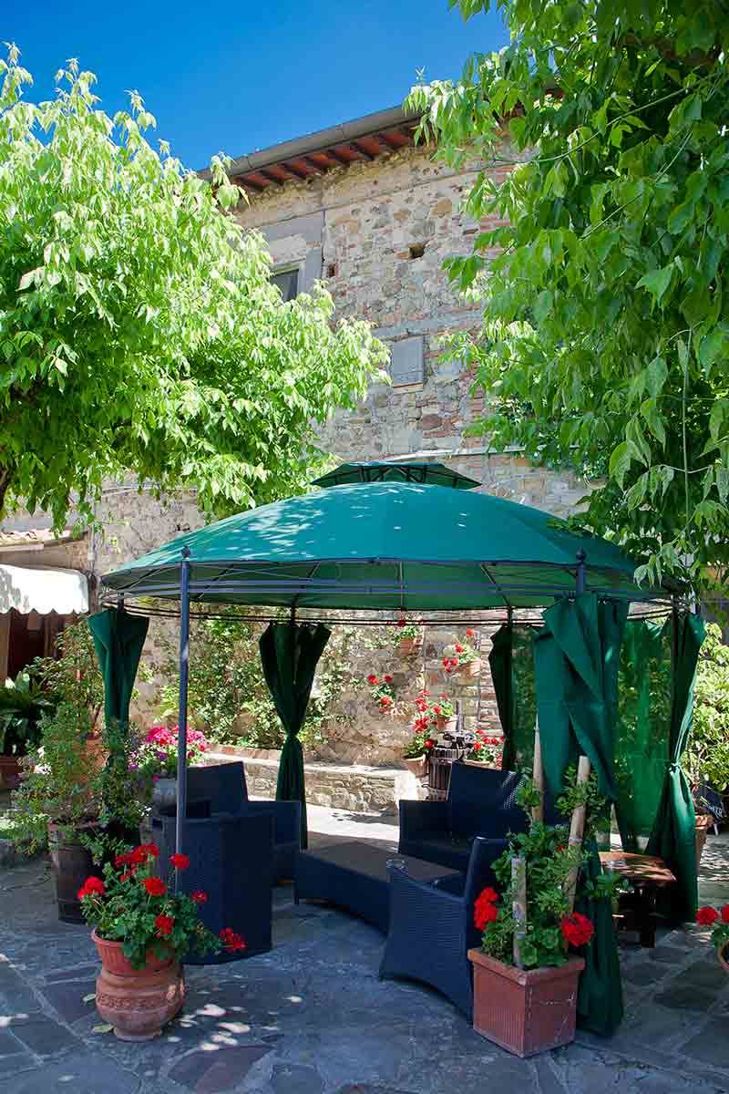 Gazebo verde con divani scuri in cortile di pietra, vasi di gerani rossi