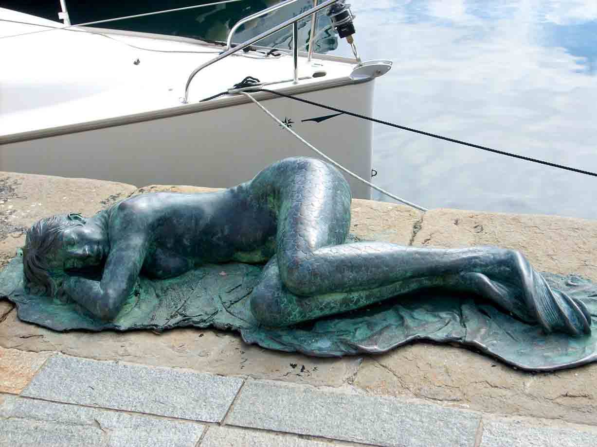 Statua bronzea di donna sdormiata sul molo, barche ormeggiate dietro