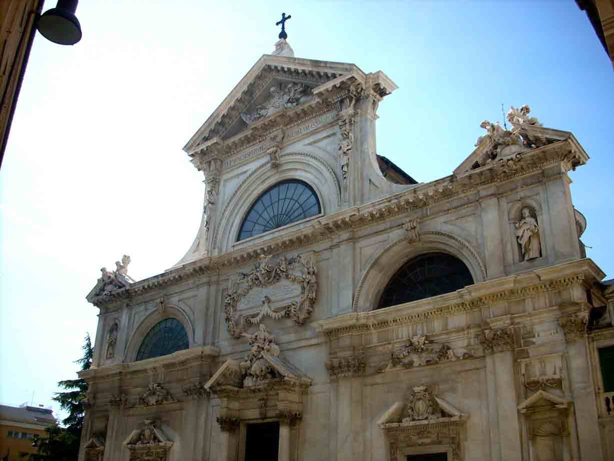 Facciata barocca in pietra chiara con statue, rosone e croce, luce solare