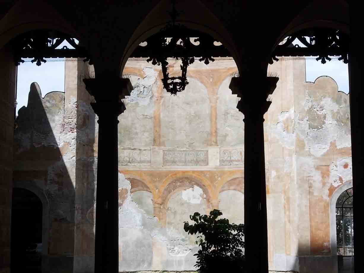 Colonne scure incontroluce su cortile con affreschi sbiaditi e pianta verde