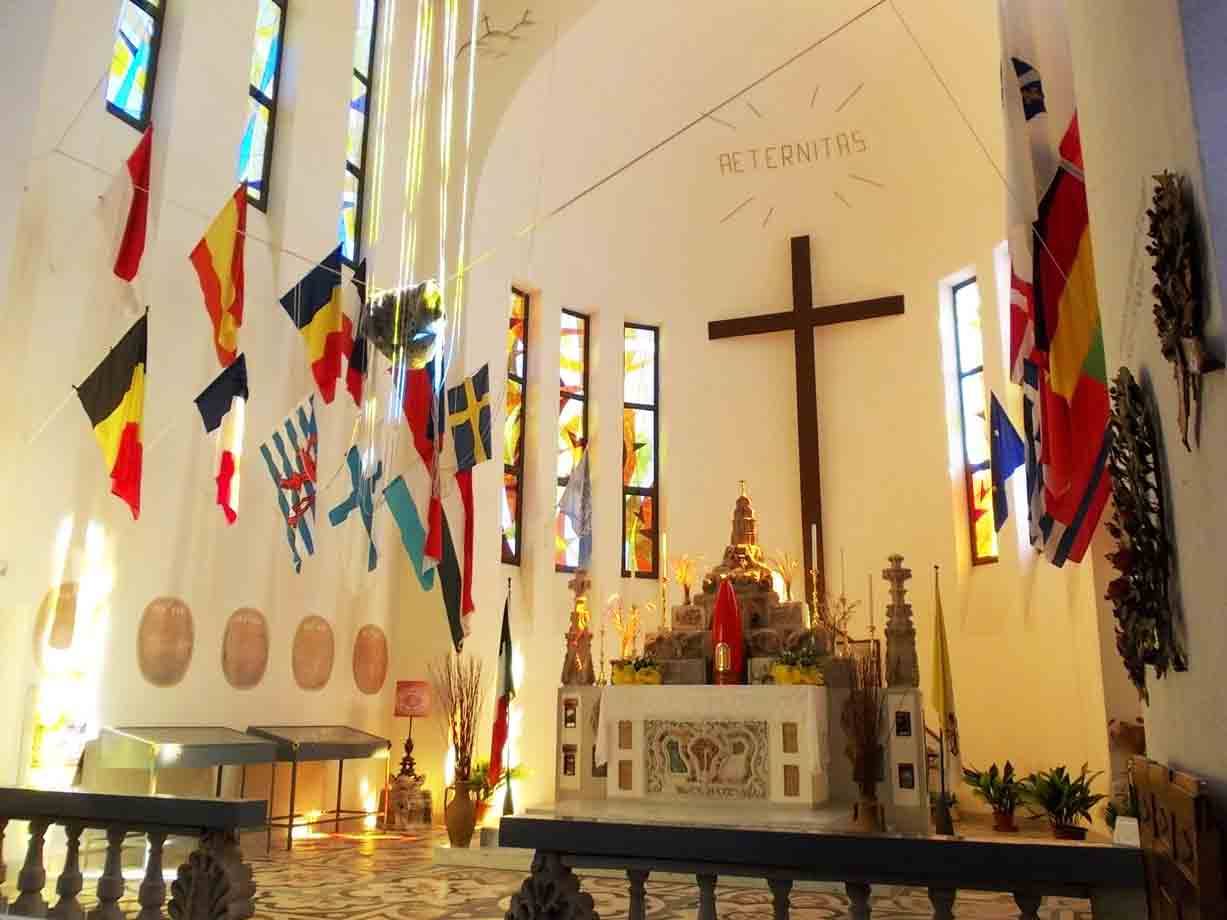 Interno di chiesa con altare, grande croce di legno e bandiere internazionali appese