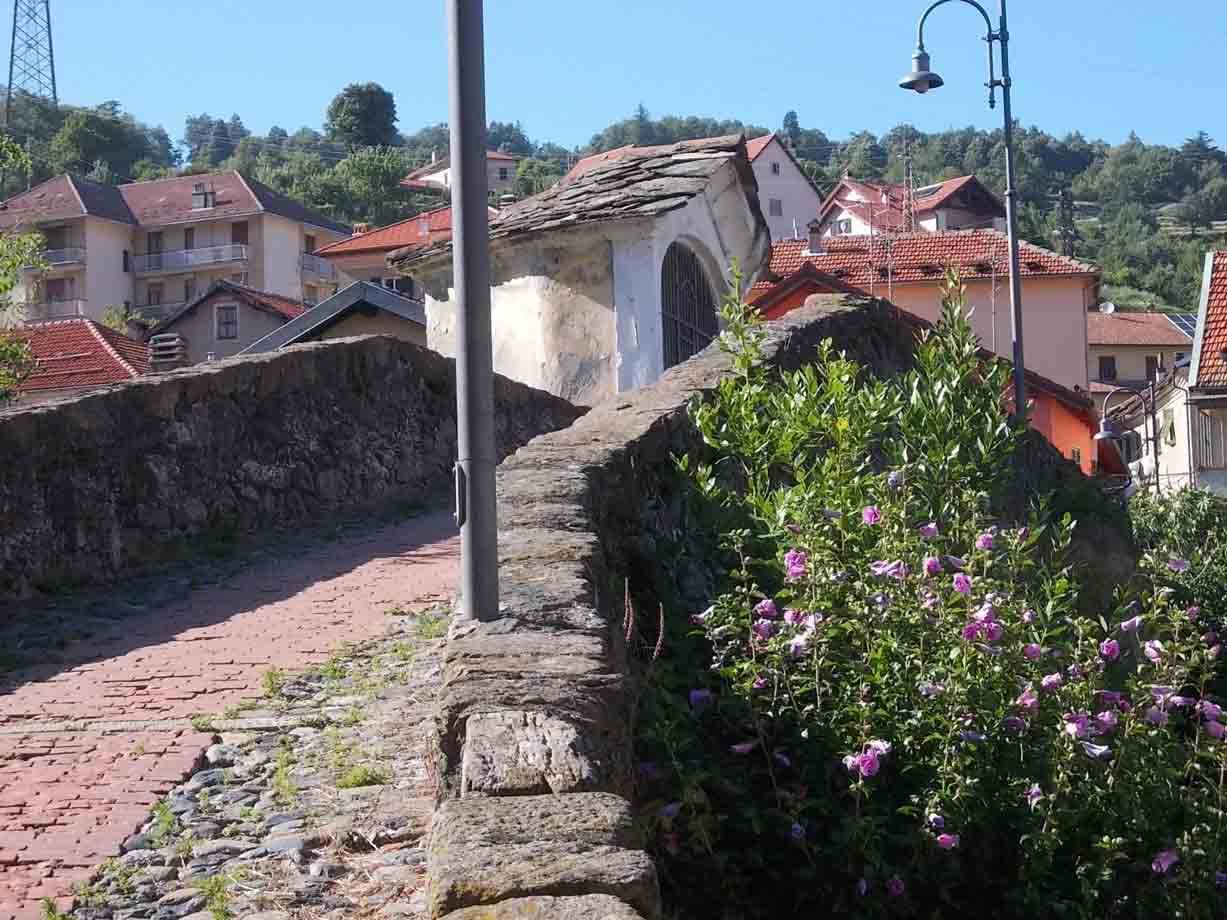 Ponte in pietra con cappella, fiori rosa e tetti rossi nel borgo