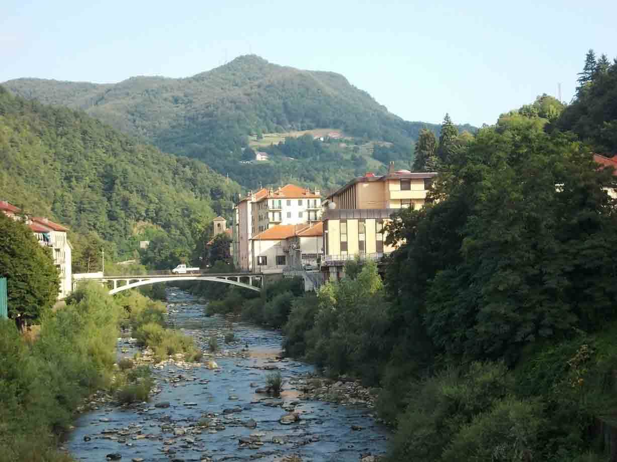 Fiume con ciottoli, ponte bianco e case tra boschi e colline verdi