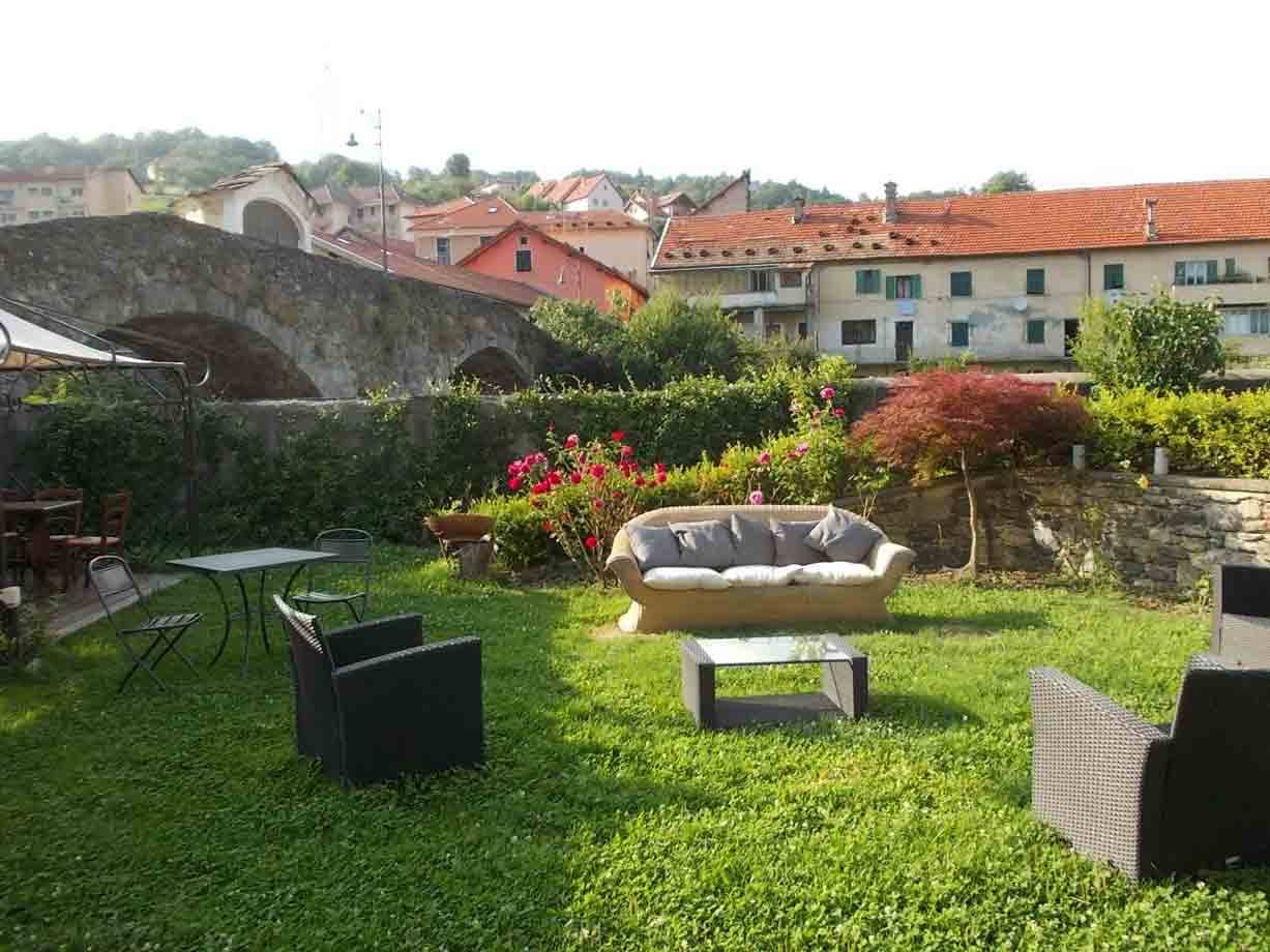 Giardino con divano e poltrone in rattan sul prato, sullo sfondo un ponte in pietra e case con tetti rossi