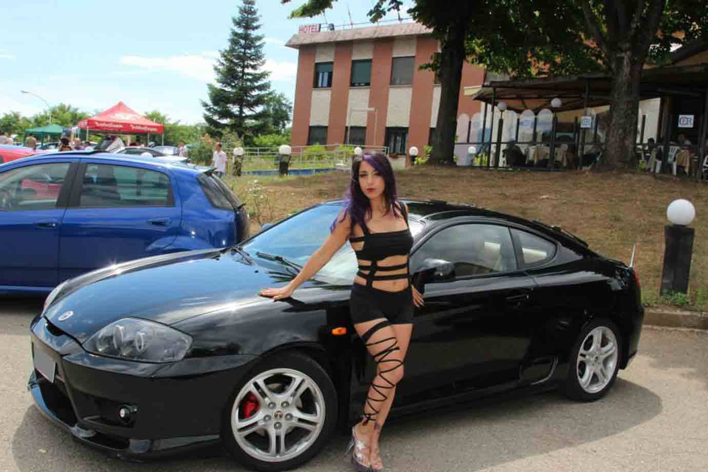 Ragazza dai capelli viola appoggiata a una Hyundai Coupé nera radunata, luce solare