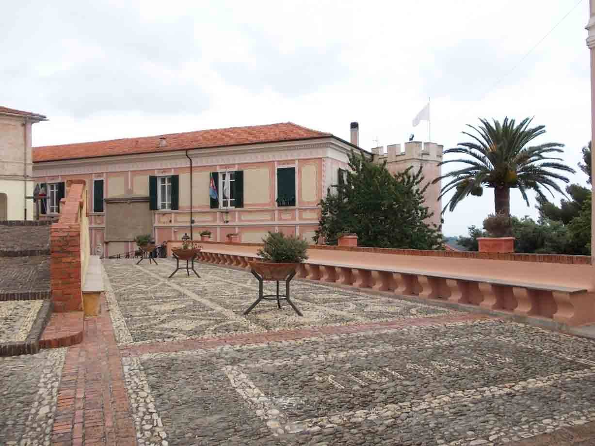 Terrazza ciottolata davanti a palazzo rosa con tetto in coppi, palme e cielo grigio