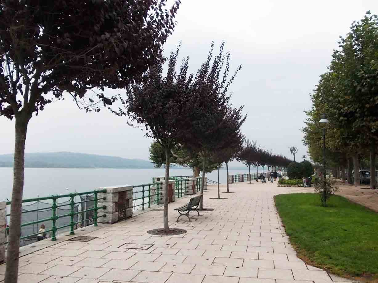 Lungolago lastricato con panchine, ringhiere verdi e alberi, persone passeggiano