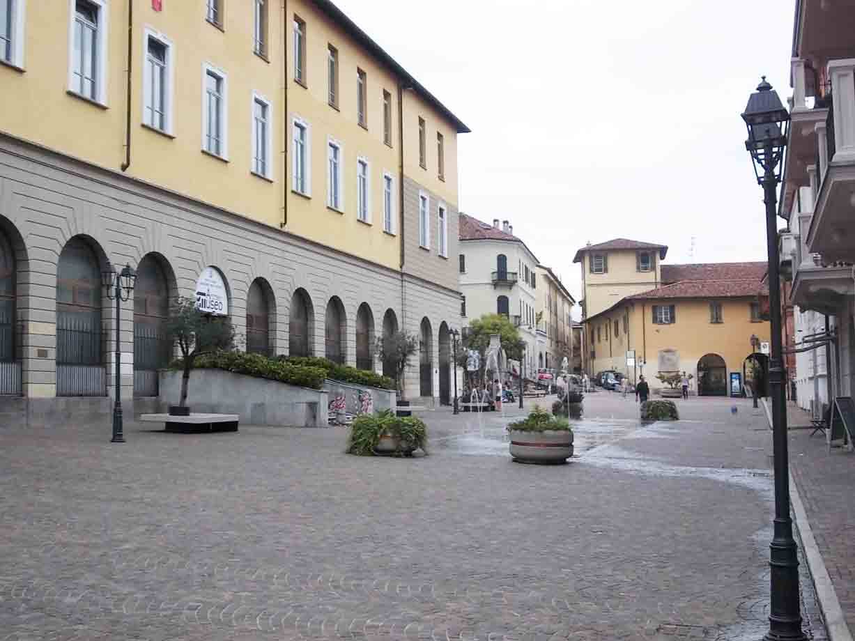 Piazza lastricata con palazzi gialli ad arcate, aiuole e lampioni sotto cielo grigio