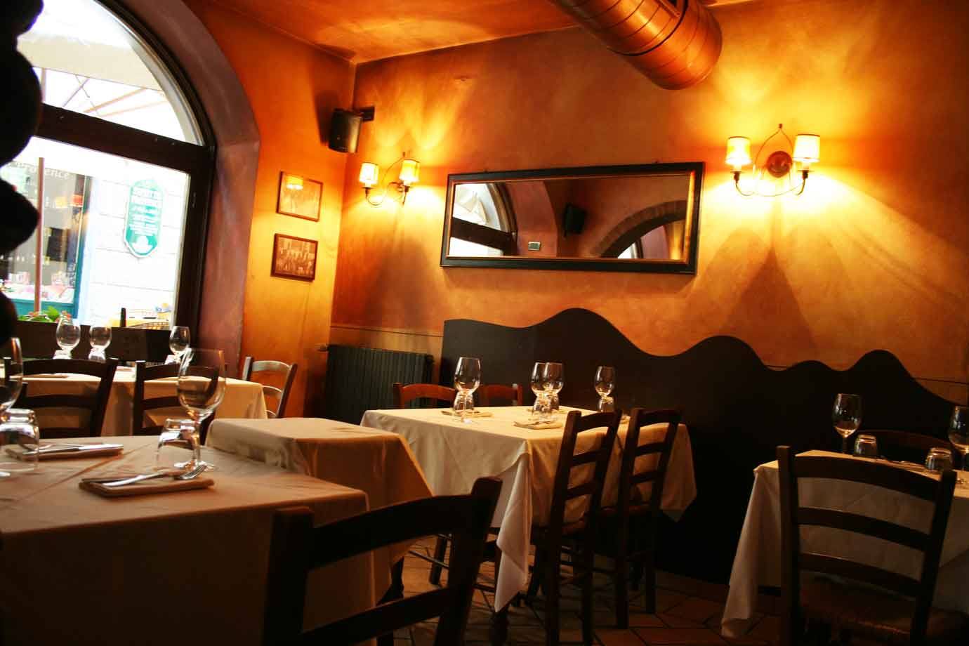 Sala di trattoria con tavoli apparecchiati, pareti ocra, luci calde a muro