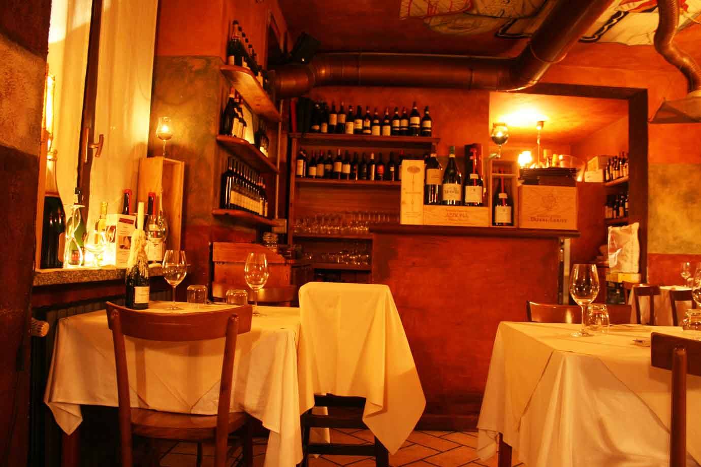Sala di trattoria con tavoli a tovaglia bianca e scaffali di bottiglie di vino