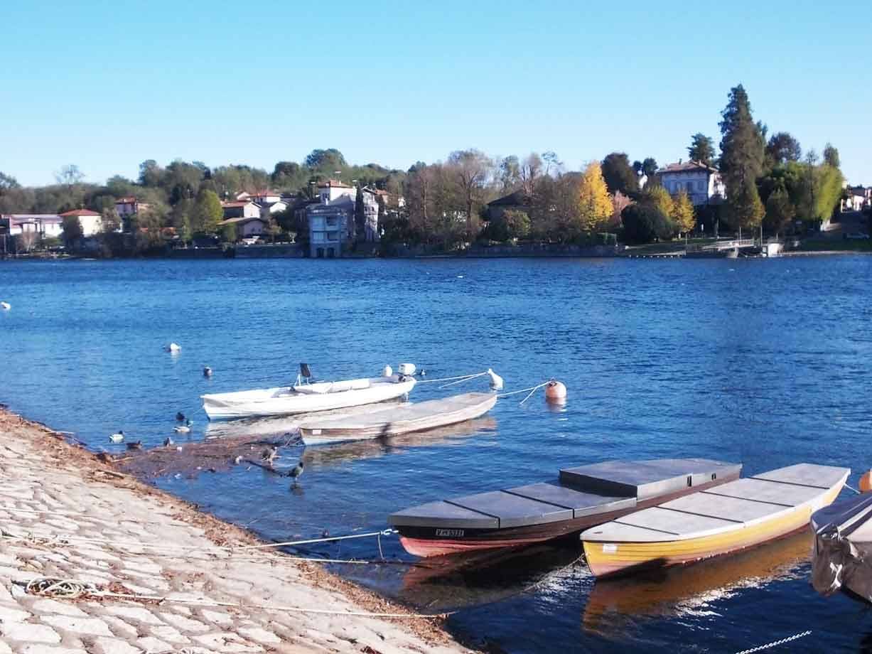 Barche a remo ormeggiate sulla riva di ciottoli, acqua azzurra e villaggio sul lago