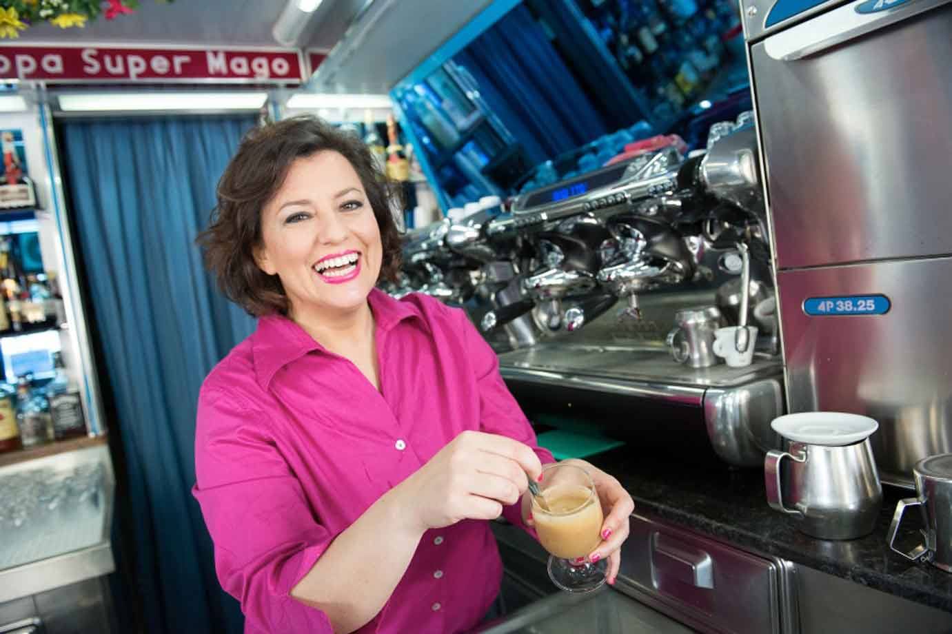 Donna sorridente in maglia fucsia mescola un caffè al bancone, macchina espresso cromata