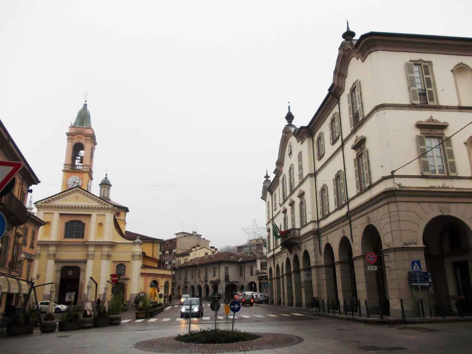 Piazza con chiesa gialla e campanile, portici e palazzo stuccato, cielo grigio