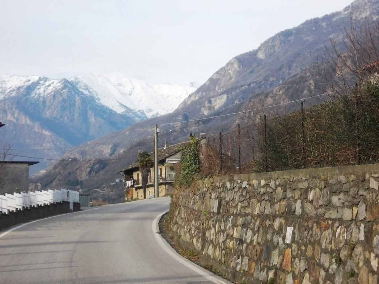 Strada di montagna con muro in pietra a secco e case tra vette innevate