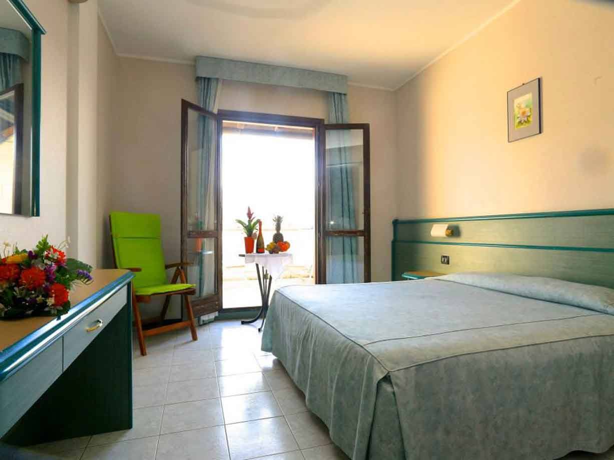 Camera d'albergo con letto azzurro, poltrona verde e porta finestra aperta