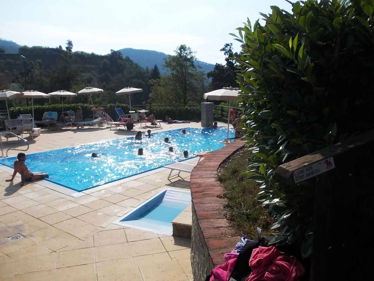 Bambini nuotano in piscina all'aperto con ombrelloni, sdraio e colline verdi sullo sfondo
