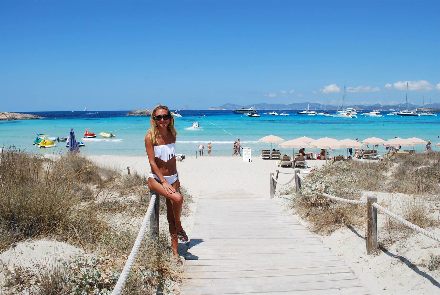 Donna in bikini bianco su passerella di legno verso la spiaggia con ombrelloni e barche