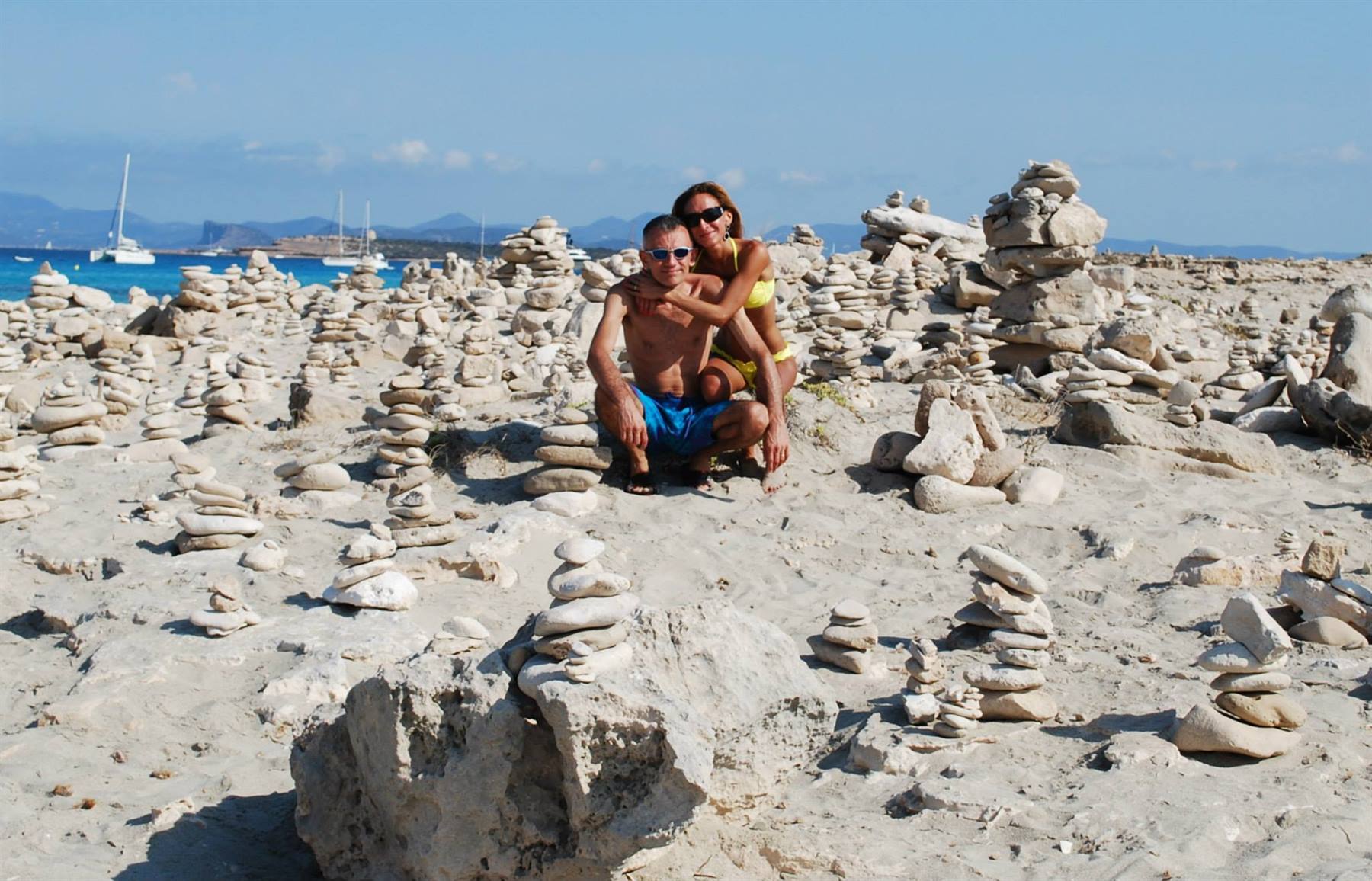 Coppia accovacciata tra mucchietti di sassi sulla spiaggia bianca, velieri al largo