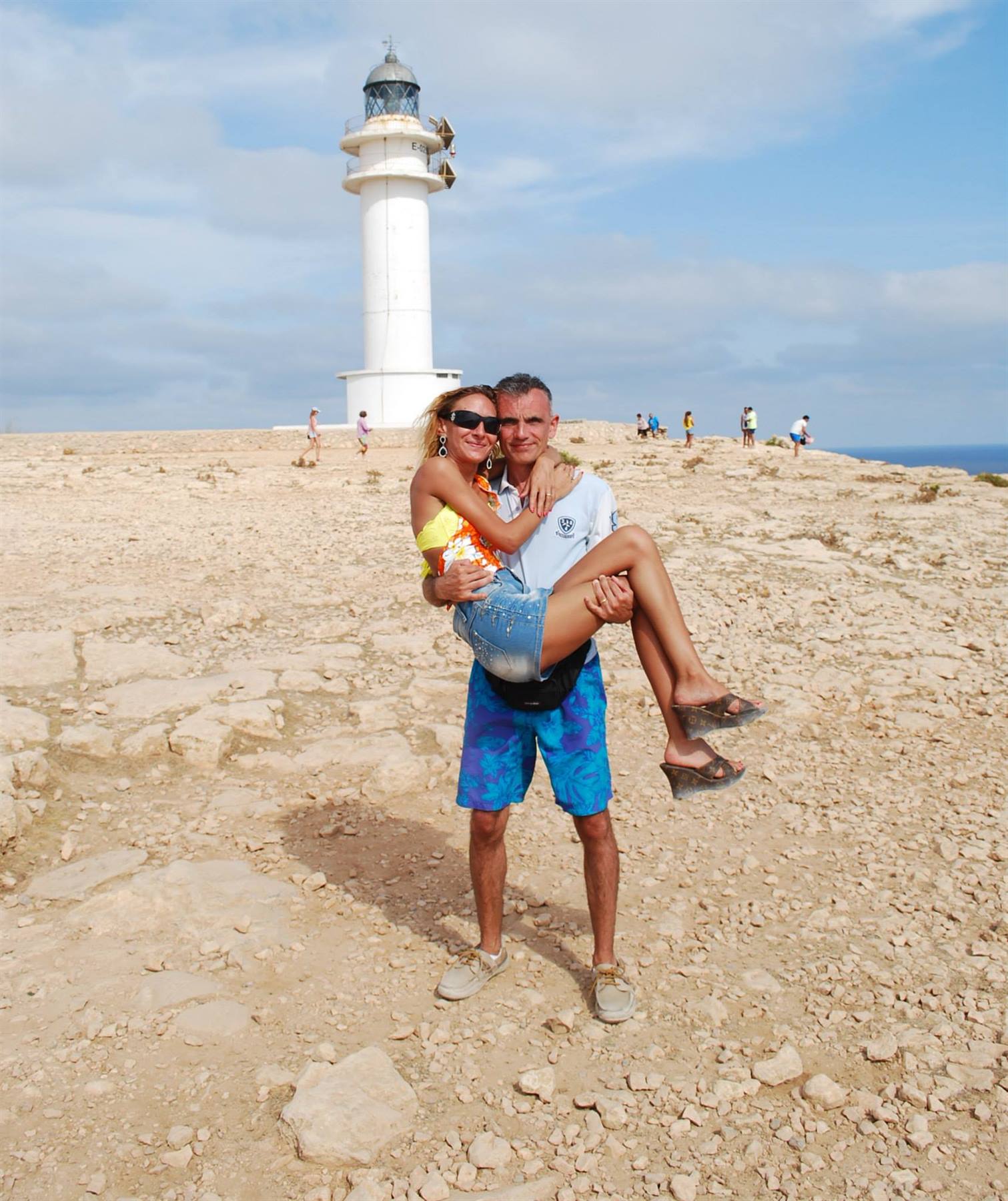 Lui la solleva in braccio davanti al faro di Barbaria su roccia arida